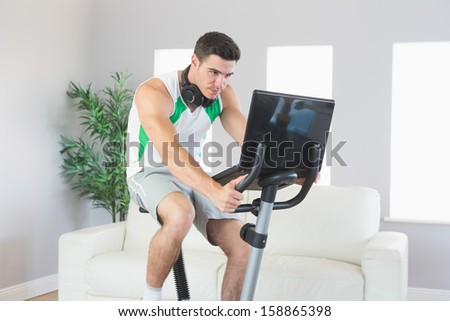 Stern handsome man training on exercise bike using laptop in bright living room