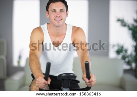 Attractive fit man exercising on bike smiling at camera in bright living room