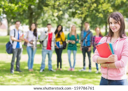 Portrait of college girl holding books with blurred students standing in the park
