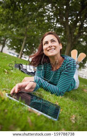 Day dreaming casual student lying on grass using tablet on campus at college