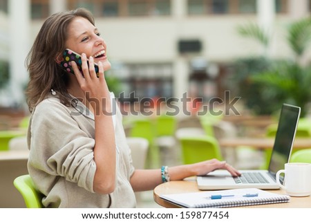 Side view of a cheerful female student using cellphone and laptop at cafeteria table