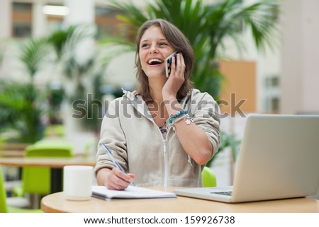Cheerful female student doing homework while on call by laptop at cafeteria table