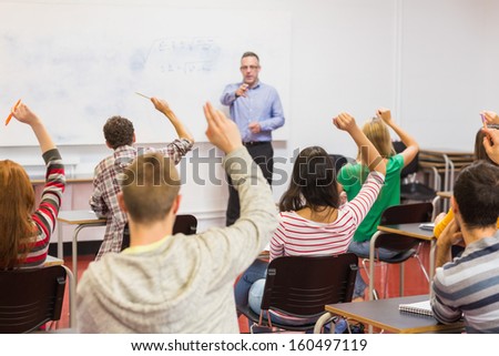 Rear view of students with hands raised with a teacher in the classroom
