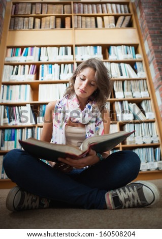 Full length of a female student sitting against bookshelf and reading a book on the library floor