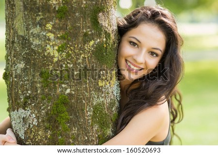 Casual smiling brunette embracing a tree looking at camera in a park on a sunny day