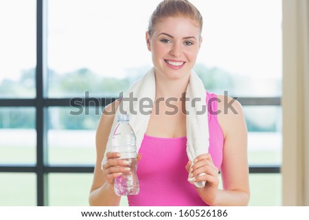 Portrait of a young woman with towel around neck holding water bottle in fitness studio