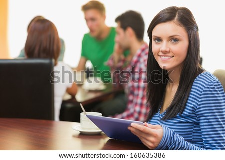 Portrait of a smiling female with coffee using tablet PC and students around table in background at  the coffee shop