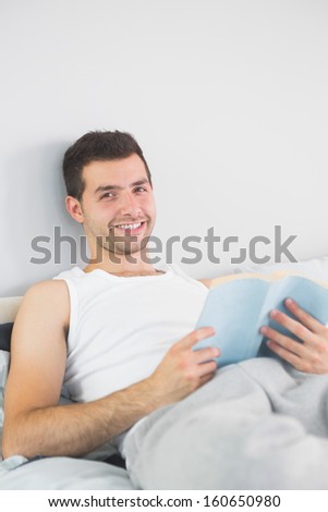 Handsome smiling man reading book in his bed in bright bedroom