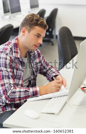 Handsome smiling student using computer taking notes in computer room at college