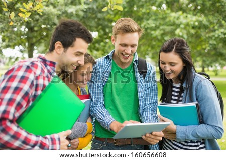 Group of cheerful college students with bags and books using tablet PC in the park