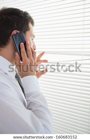 Attractive businessman spying through roller blind while phoning in bright office