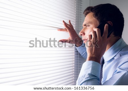 Side view of a young businessman peeking through blinds while on call in office