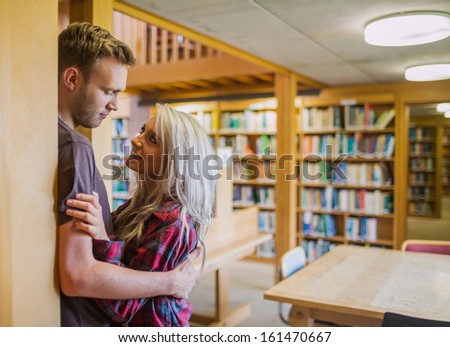 Side view of a young romantic couple looking at each other with bookshelf at a distance in the library