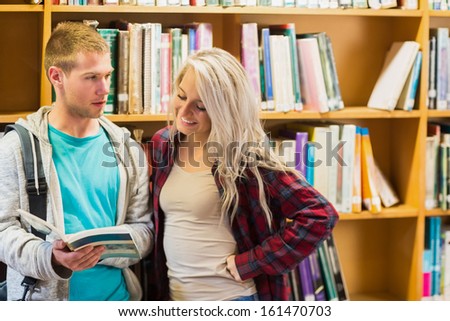 Two young students reading book against bookshelves in the library
