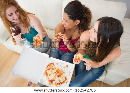 Overhead view of happy young female friends eating pizza with wine on sofa at home