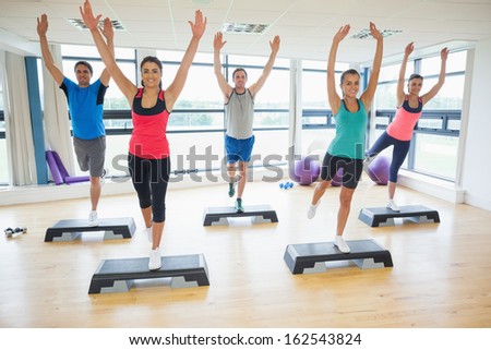 Full length portrait of instructor with fitness class performing step aerobics exercise in gym