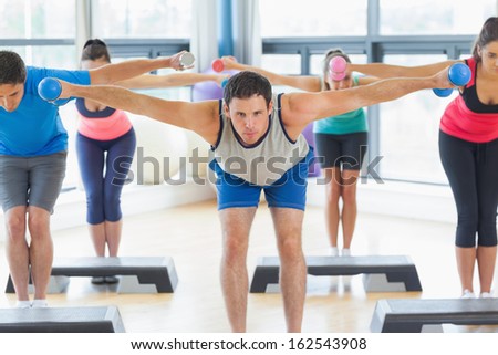 Full length of instructor with fitness class performing step aerobics exercise with dumbbells in a gym
