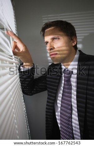 Handsome serious young businessman peeking through blinds in the office