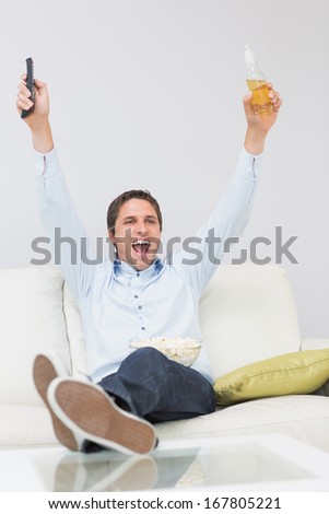 Cheerful young man with a drink and remote control cheering on sofa in the living room at home