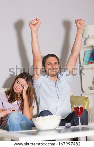Excited man watching TV with wine glass and popcorn besides a bored woman on sofa at home