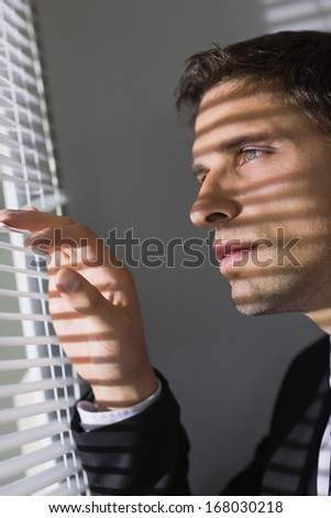 Close-up side view of a serious young businessman peeking through blinds in the office