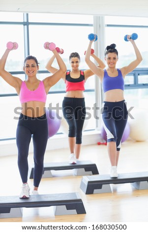 Full length fitness class performing step aerobics exercise with dumbbells in a gym