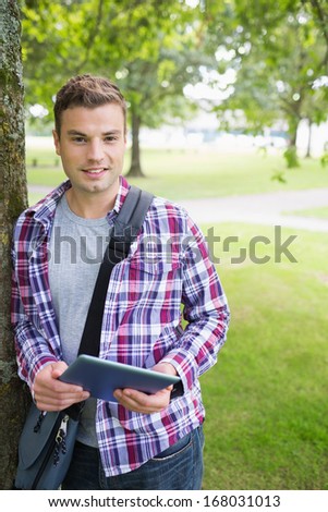 Handsome student leaning on tree looking at camera holding tablet on college campus