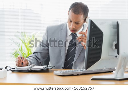 Concentrated young businessman using computer and phone at office desk