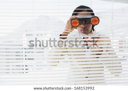 Serious young businessman peeking with binoculars through blinds in the office
