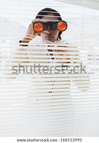 Serious young businesswoman peeking with binoculars through blinds in the office