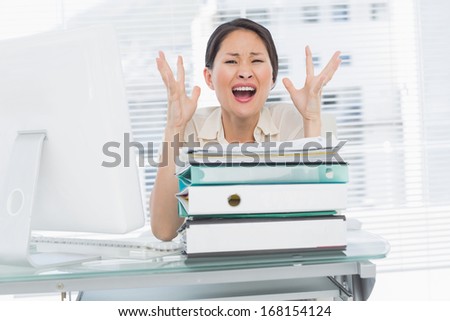 Portrait of a young angry businesswoman shouting with stack of folders at office desk