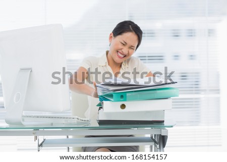 Angry young businesswoman sitting with stack of folders at office desk