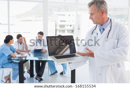 Doctor holding laptop with group around table in background at hospital