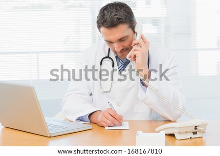 Concentrated male doctor using phone while writing notes by laptop at the medical office