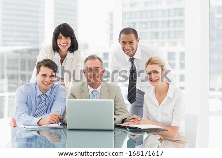 Happy business people gathered around laptop looking at camera in the office