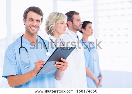 Portrait of a male doctor writing reports with colleagues standing behind in a medical office