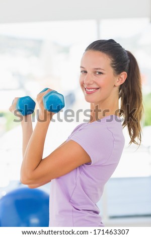 Side view of a smiling young woman lifting dumbbell weights in a bright gym