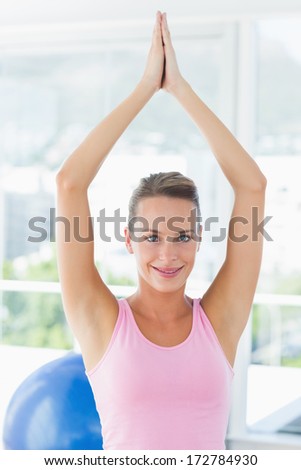 Portrait of a smiling young woman with joined hands over head at a bright fitness studio