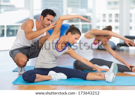 Women doing stretching exercises as trainer helps one at fitness studio