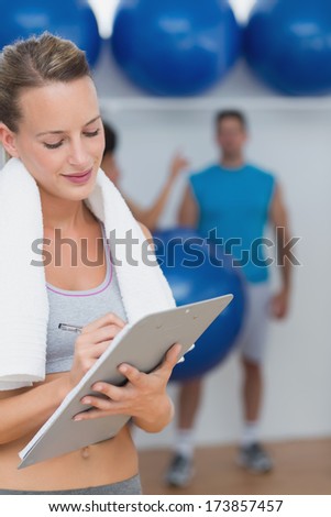 Female trainer writing on clipboard with fitness class in background at the gym