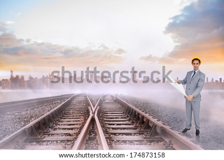 Serious architect with hard hat holding plans against train tracks leading to misty city on the horizon