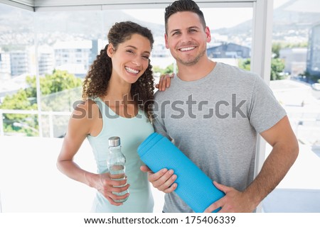 Portrait of fit couple holding water bottle and exercise mat in bright exercise room