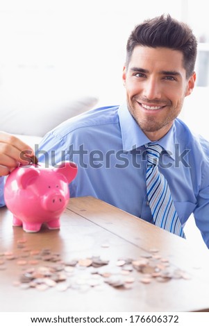 Handsome businessman putting coins into piggy bank looking at camera in the office