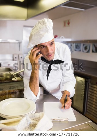 Concentrated male cook writing on clipboard while using cellphone in the kitchen