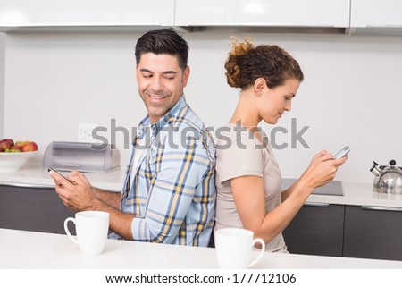 Happy couple sitting back to back texting at home in kitchen