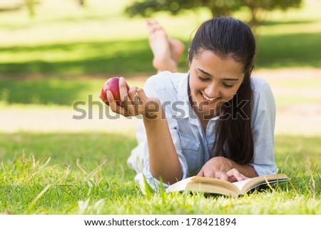 Smiling young woman eating apple while reading a book in the park