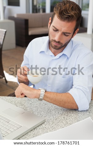 Frowning young businessman checking time holding coffee cup in patio of restaurant