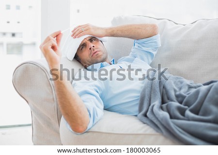 Sick man lying on sofa checking his temperature under a blanket at home in the living room