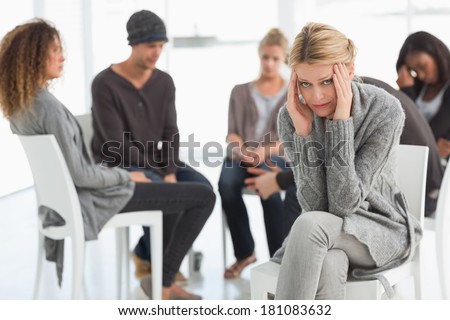Upset woman with head in hands at rehab group looking at camera at therapy session