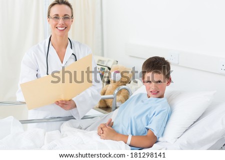 Portrait of happy female doctor holding report with sick boy in hospital bed